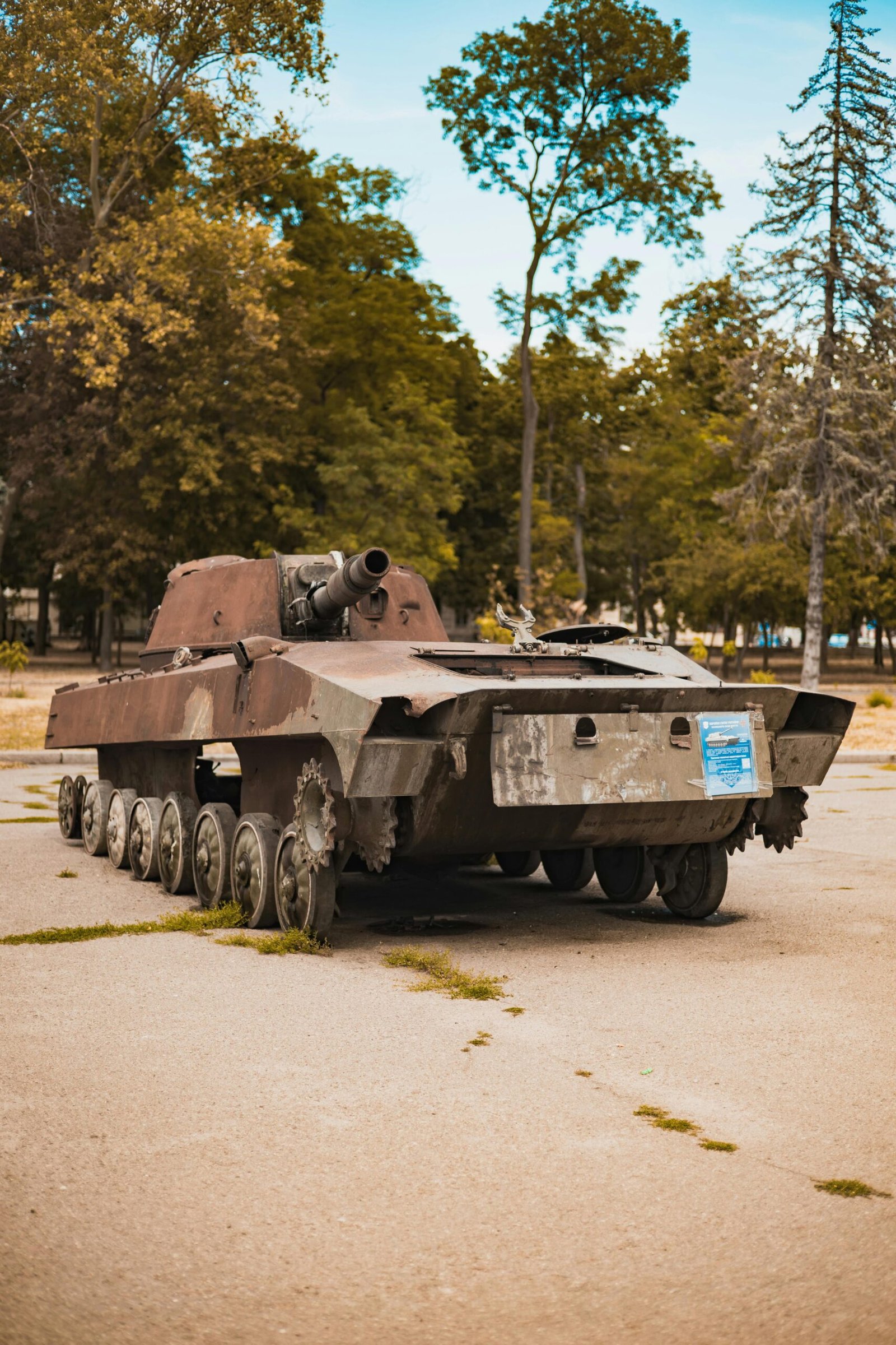 An old tank is abandoned in a park setting surrounded by trees and clear sky.